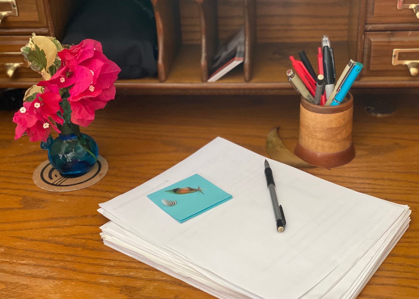 A sheaf of manuscript pages on a wooden desk with pencil, vase of flowers, and a round carved wooden pen and pencil holder. On the sheaf of papers is a nearly used-up stack of Post-It notes and two tiny feathers