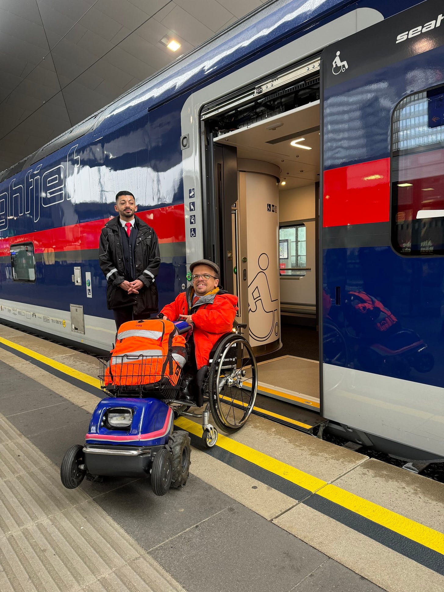 Wheelchair user Andreas Pöschek together with a train manager at the open door of the new nightjet. Andreas has a small wheelchair with an electric attachement at the front and a big orange bag on top of it. The entrance is flat with a small ramp.