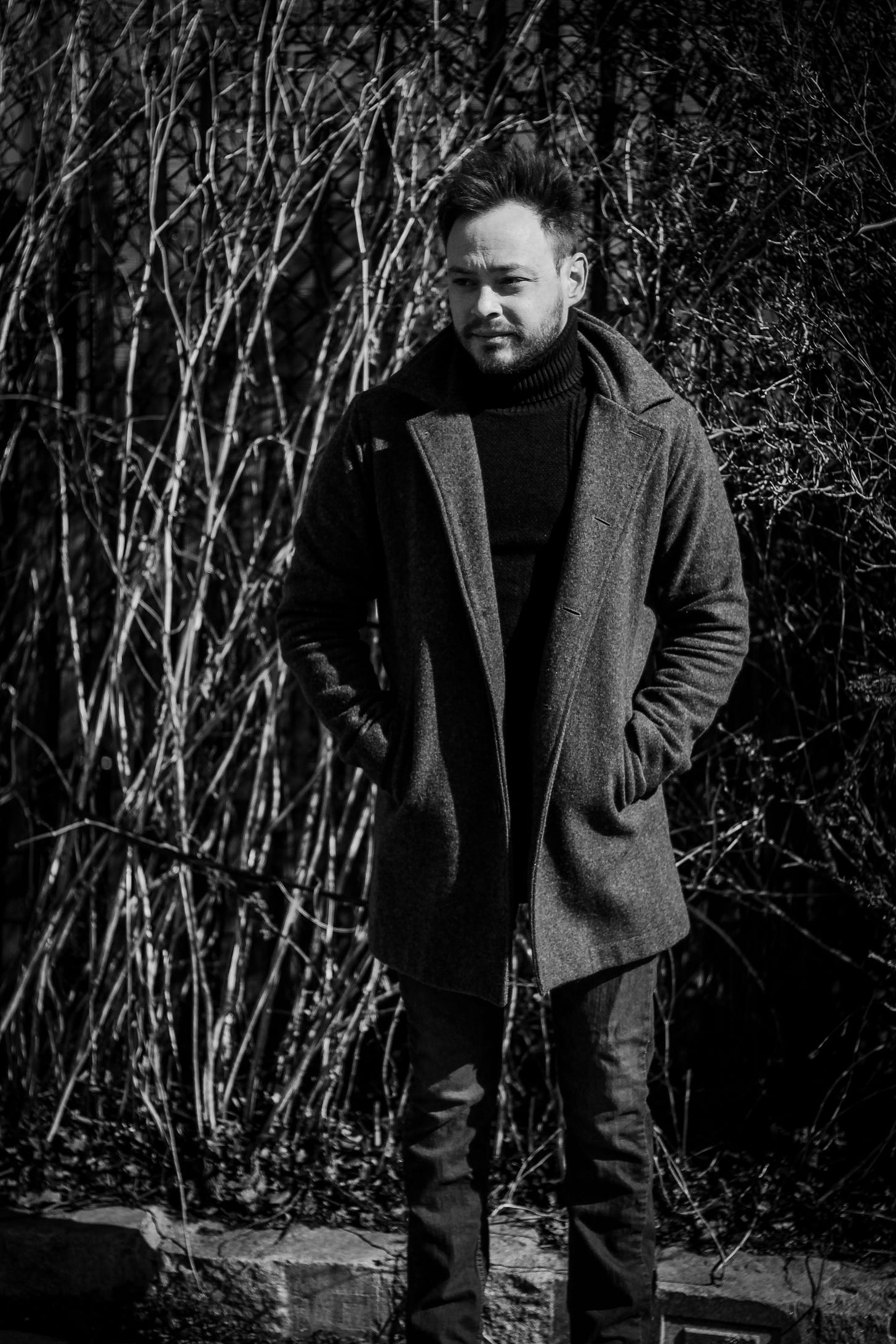 Photo of composer Charlie Rauh standing in a forest in a coat with hands in pockets.