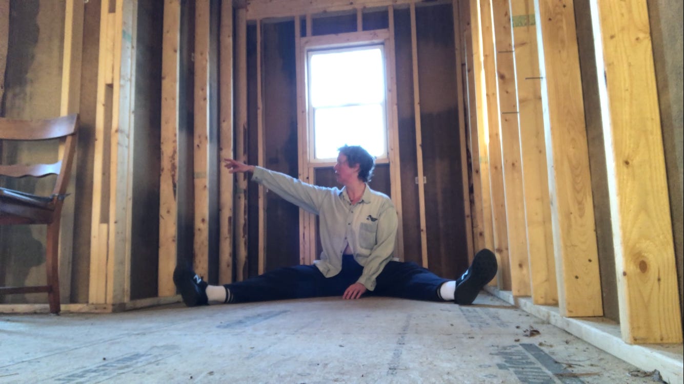 a person sitting on the floor of an unfinished building reaching and looking to the left, legs splayed
