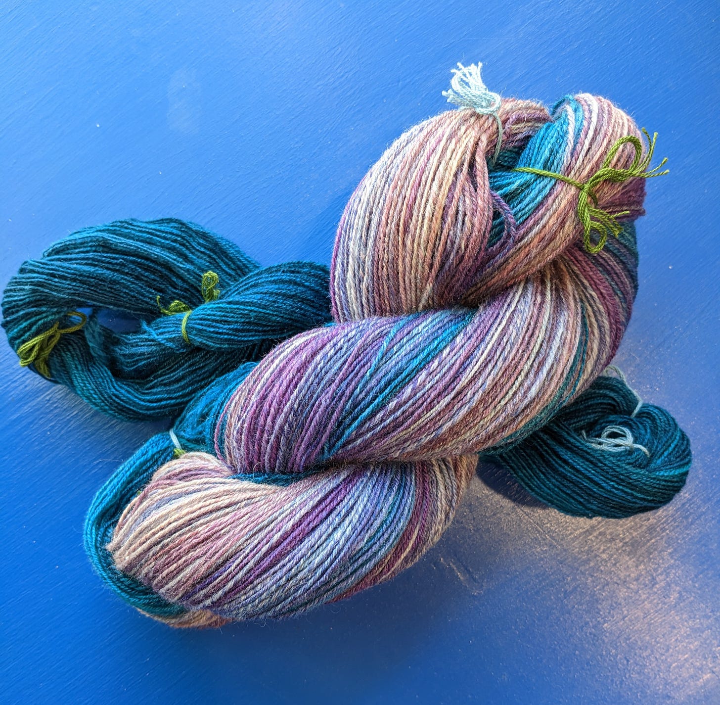 Two skeins of yarn (one dark teal, one an ombre from dark teal to peachy purple tones) on a blue background.