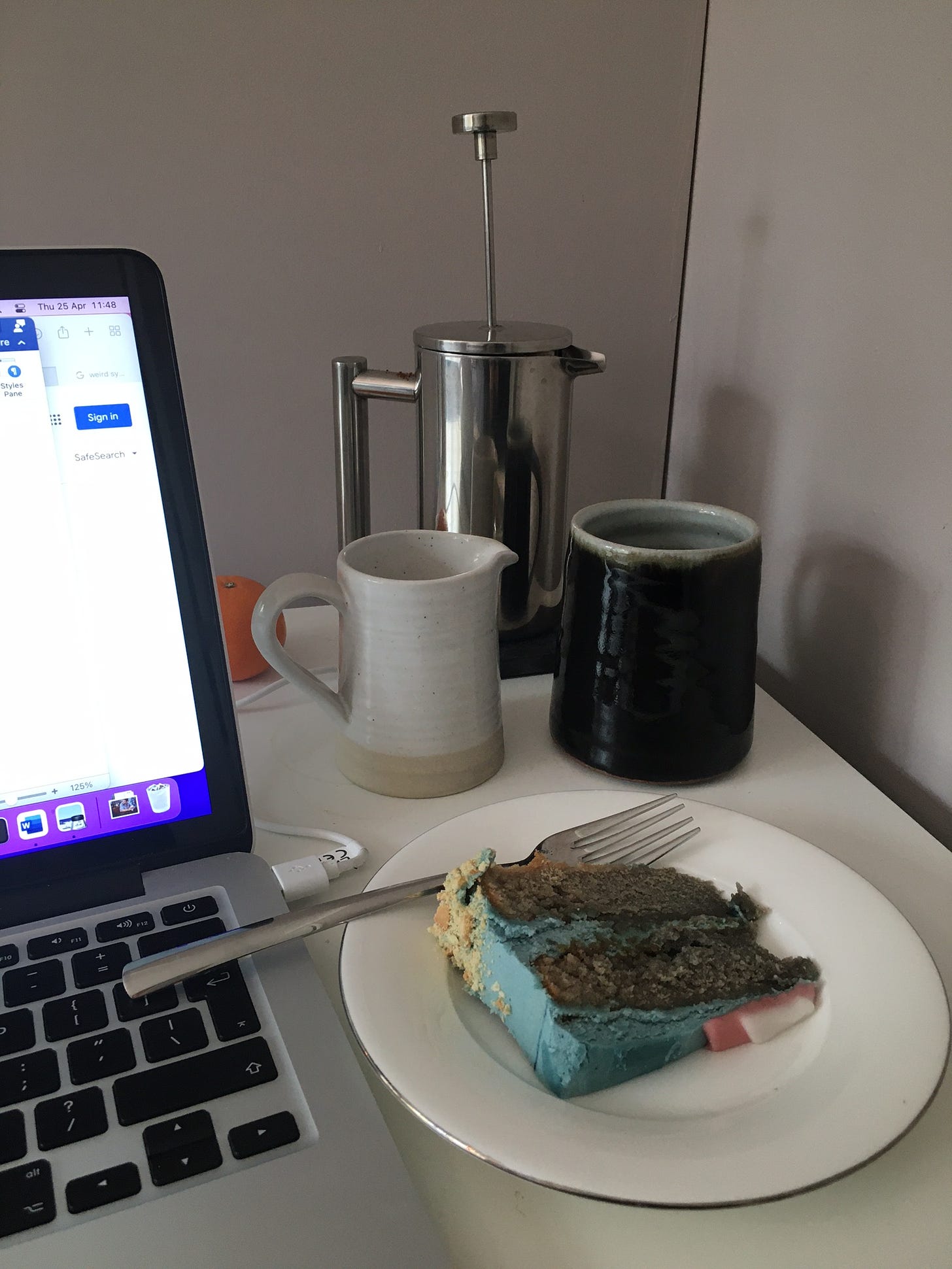 piece of cake with blue icing, coffee cup, milk jug and cafetière next to an open laptop