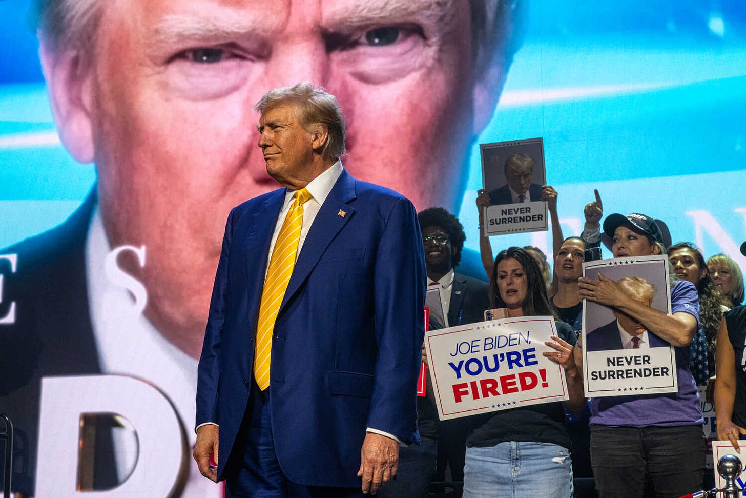 Donald Trump, wearing a blue suit and a yellow striped tie, stands onstage in front of a large projection of his face and supporters holding signs that read “Joe Biden You’re Fired” and “Never Surrender.”