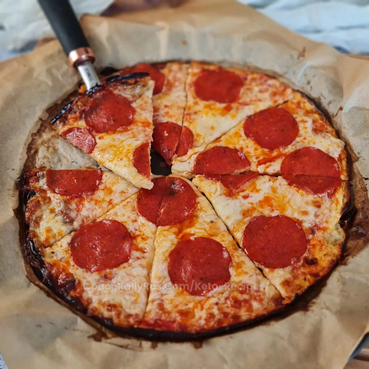 A freshly baked pizza with a golden-brown crust and melted cheese, topped with large pepperoni slices. The pizza is cut into triangular slices and rests on parchment paper with a pizza cutter on top, suggesting it is a homemade or artisanal cottage cheese pizza.