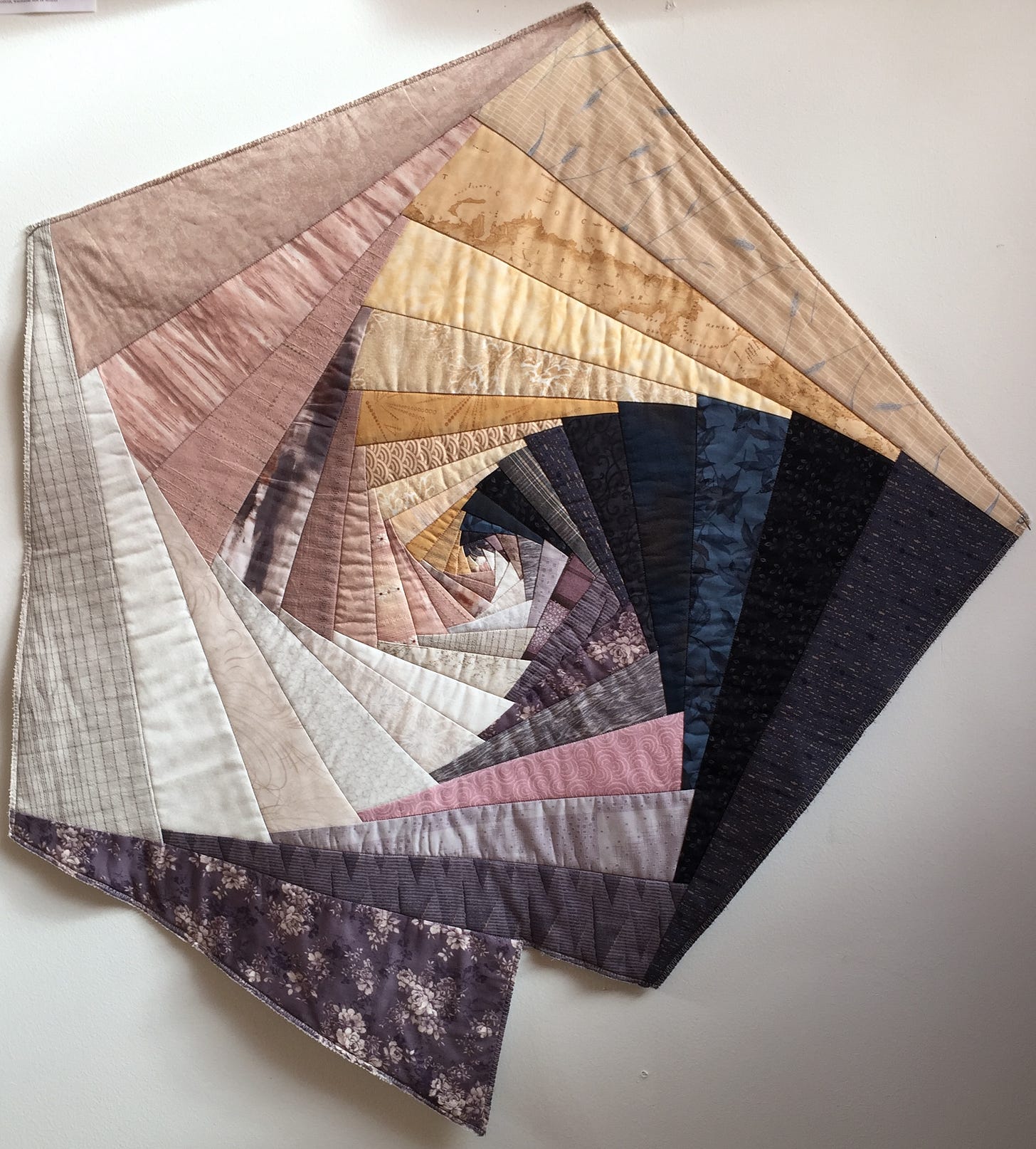 The work is a free-form pieced quilt, an abstract imitation of a whelk shell, weaving together inside and outside. Pale gold, mauve, blue, purple, and cream fabrics entwine in a spiraling form, emerging in small pieces from the shell's center to larger and larger pieces at the shell's five-sided outer edges.