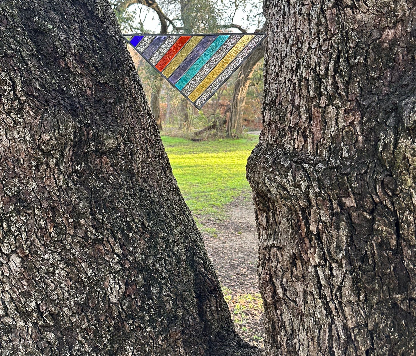 A stained glass rainbow triangle rests in the middle of a gap in tree outside.