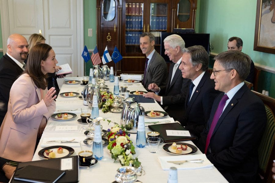 Secretary Blinken meets with Finnish Prime Minister Sanna Marin, while sitting at a long meeting table with other U.S. and Finnish officials. At the head of the table stand small NATO, U.S., Finland, and EU flags.