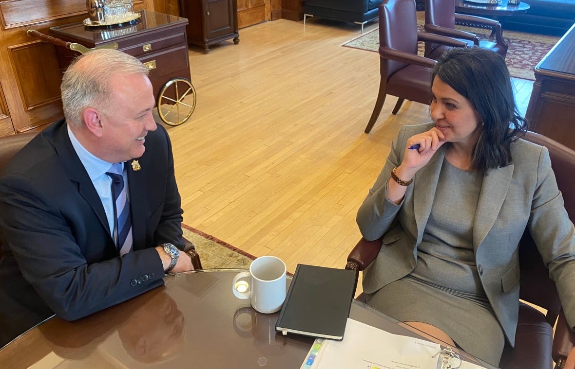 Premier Danielle Smith, on the right, sits leaning back from an office table. On her left, a man in a suit looks at her and smiles. 