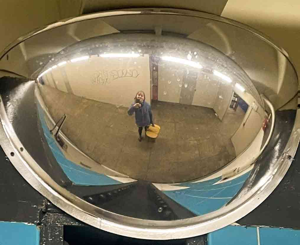 A girl takes a selfie in the reflection of a corner mirror in a subway station. She wears a purple rain jacket and holds a large green picnic basket with a wooden top.