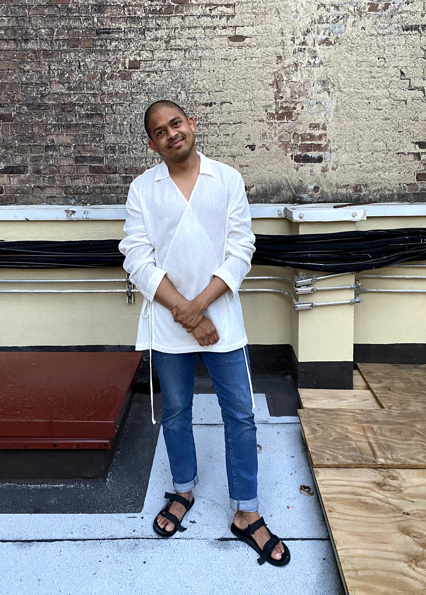 Kirk Fernandes standing in white shirt and sandals posing in front of a brick wall.
