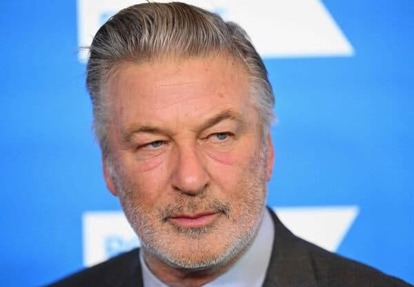 A close-up image of Alec Baldwin wearing a suit in front of a bright blue background.