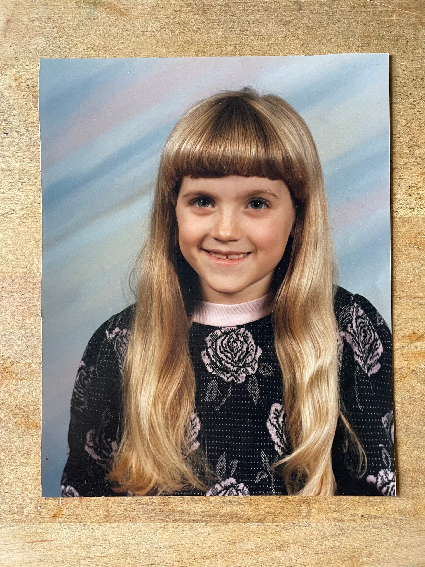 on a wooden table there is an olan mills style photograph with a purple pink background. there is a child about 8 or 9 with thick blonde bangs and hair and a gaap toothed smile. The eyes are bright but also deep. 