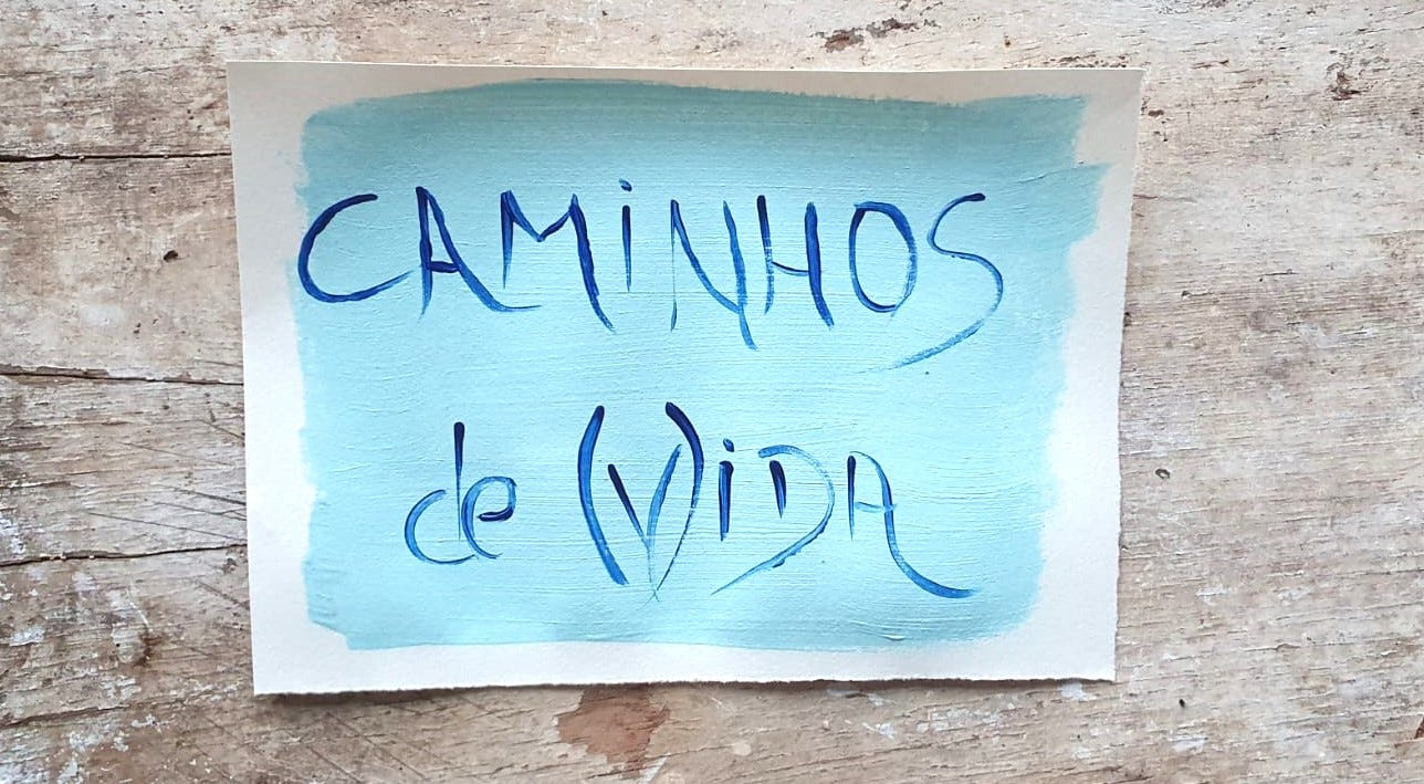 Fotografia de um papel pintado de azul claro, com a frase Caminhos de (v)ida escrita, sobre um fundo de madeira 
