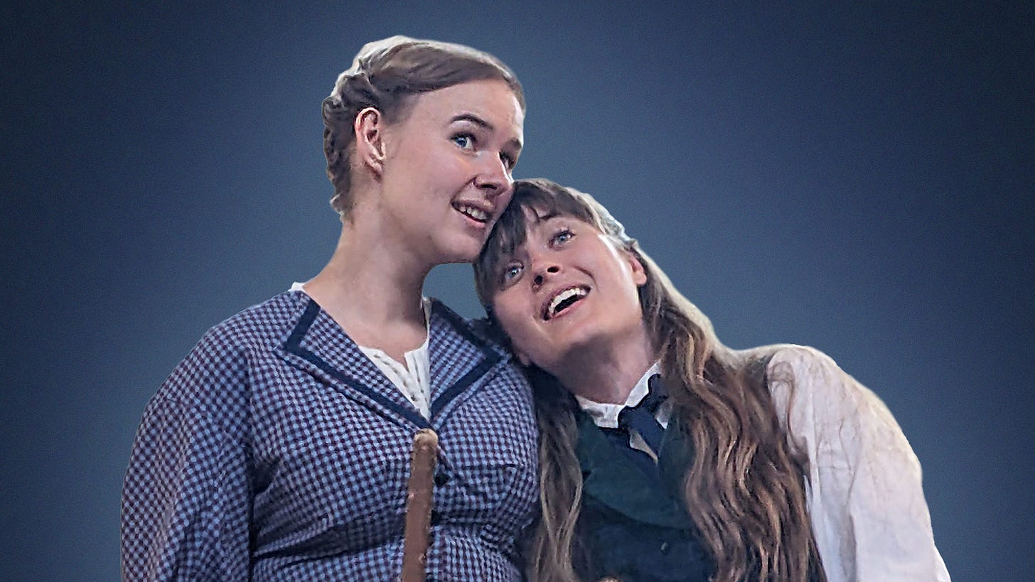 Meg and Jo, the two elder March sisters, enjoy a hug. Meg is dressed in blue gingham, Jo wears a man's collar and tie with long hair.