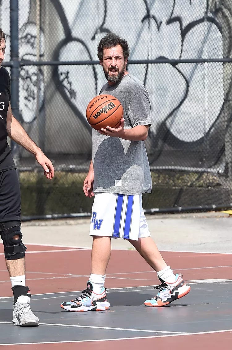 Adam Sandler in big shorts (but not the biggest) on a basketball court.