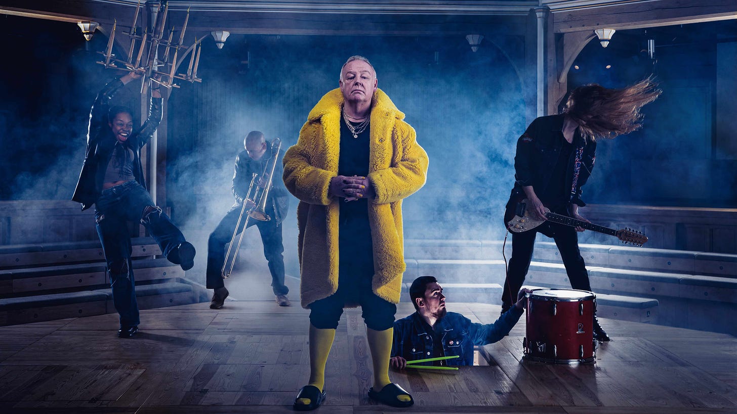 British comedian Les Dennis, wearing a woolly yellow coat and yellow stockings, stairs forward whilst actor-musicians play modern instruments on stage behind him.