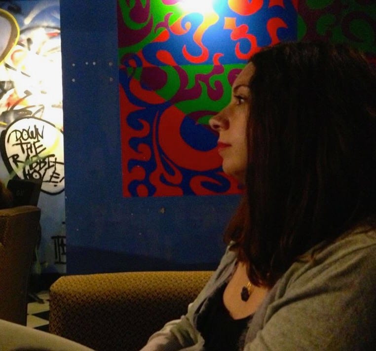 A person with shoulder-length black hair and dark eyes sits in a coffeeshop, listening. There is a bright abstract painting on the blue wall behind them.