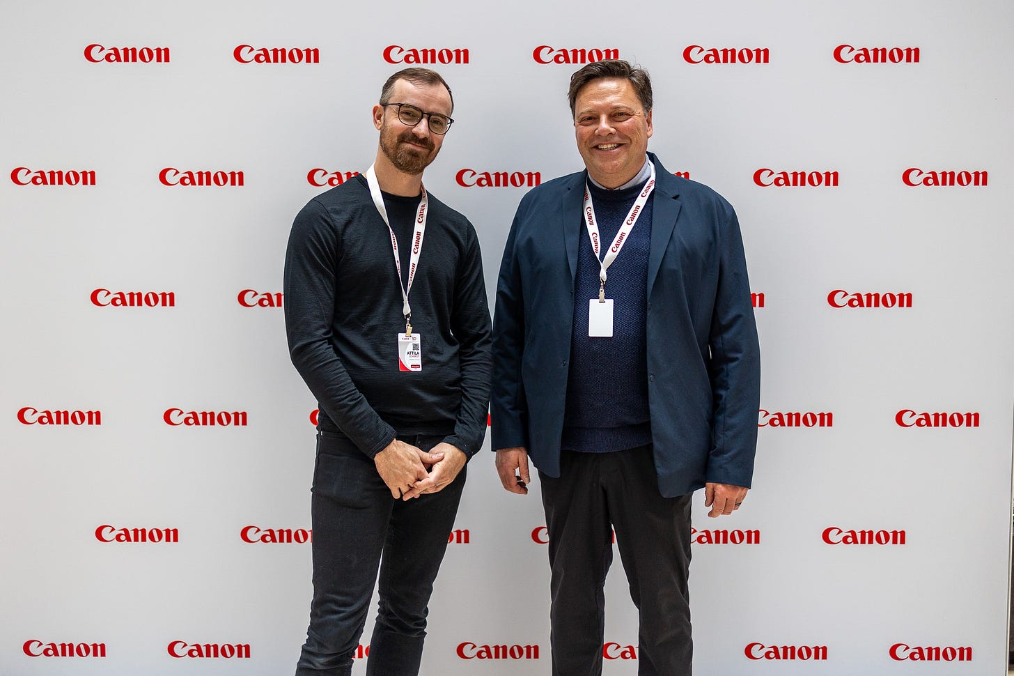 The photograph is of two people standing in front of a Canon background with the logo for Canon repeated multiple times. On the left is Attila Schmidt, wearing a black shirt and black pants. On the right is Wes Worsfold, wearing a blue jacket over a black shirt and black pants. They are both smiling.