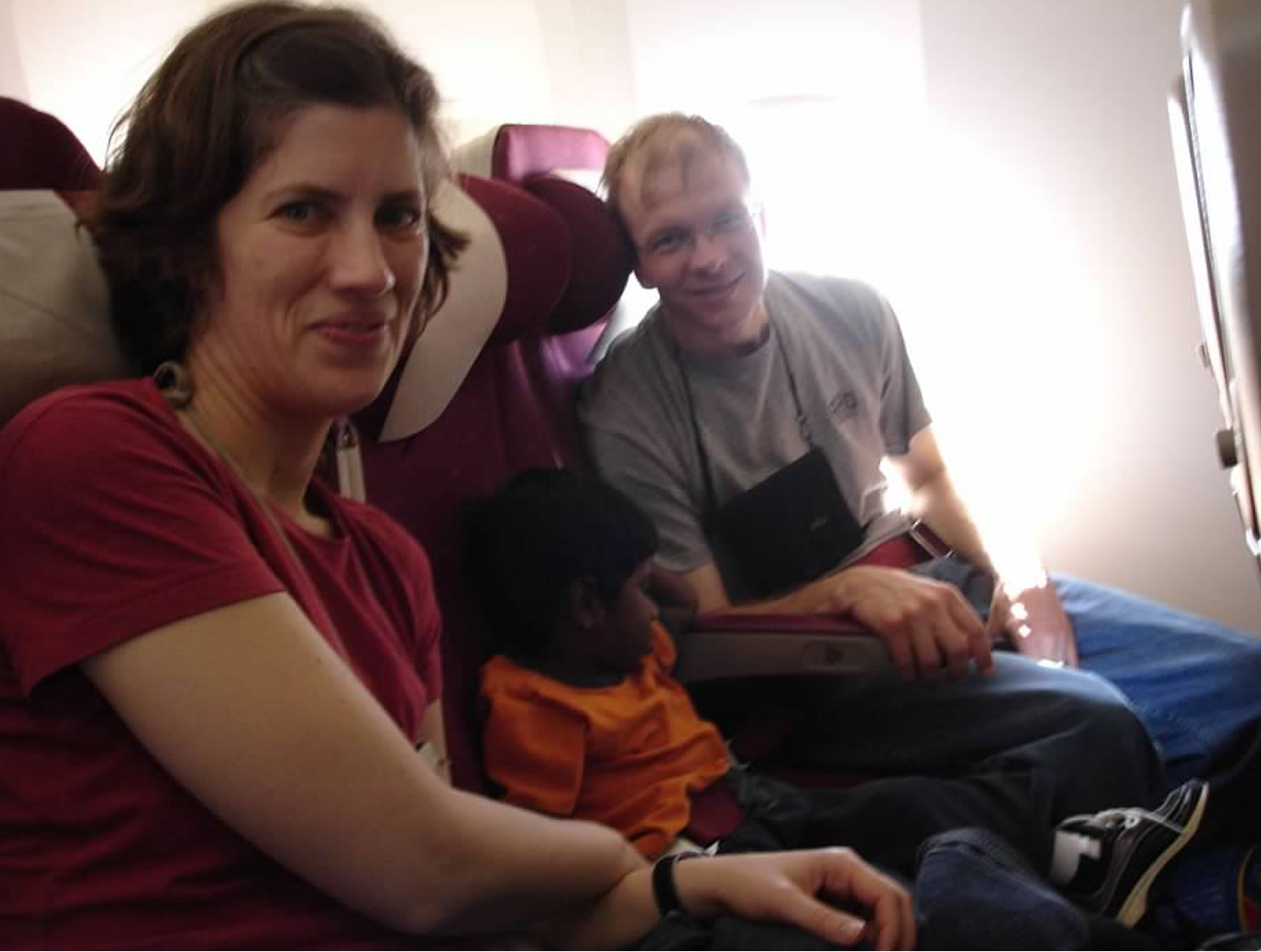 A family seated in airplane seats, the man and woman smiling at the camera with a sleeping child between them.