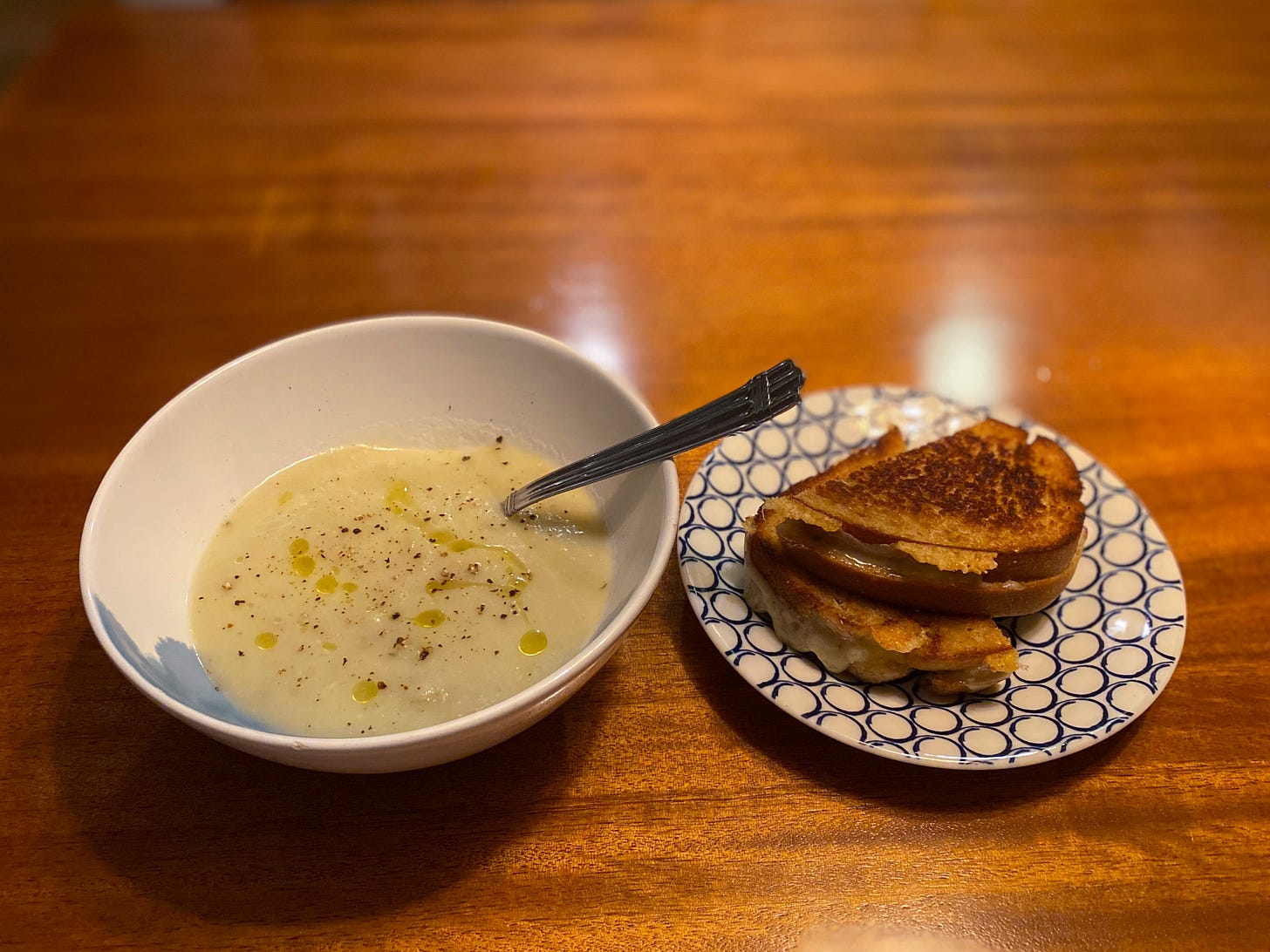 A white and blue bowl of potato-leek soup, sprinkled with pepper and drizzled with olive oil, next to a white and blue side plate with a grilled cheese on it. The sandwich is cut in half with melted cheese coming out the edges, burned and crisped in places where it's touched the pan.