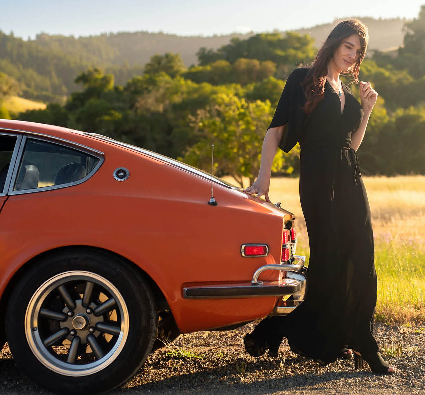 Cara Esten, a transgender woman wearing a long flowing black dress, poses with her bright orange Datsun 240Z, her hand resting on the back trunk lid from the vehicle's side profile against a sunny open meadow in the background. For the new book 'We Deserve This' by Victoria Scott.