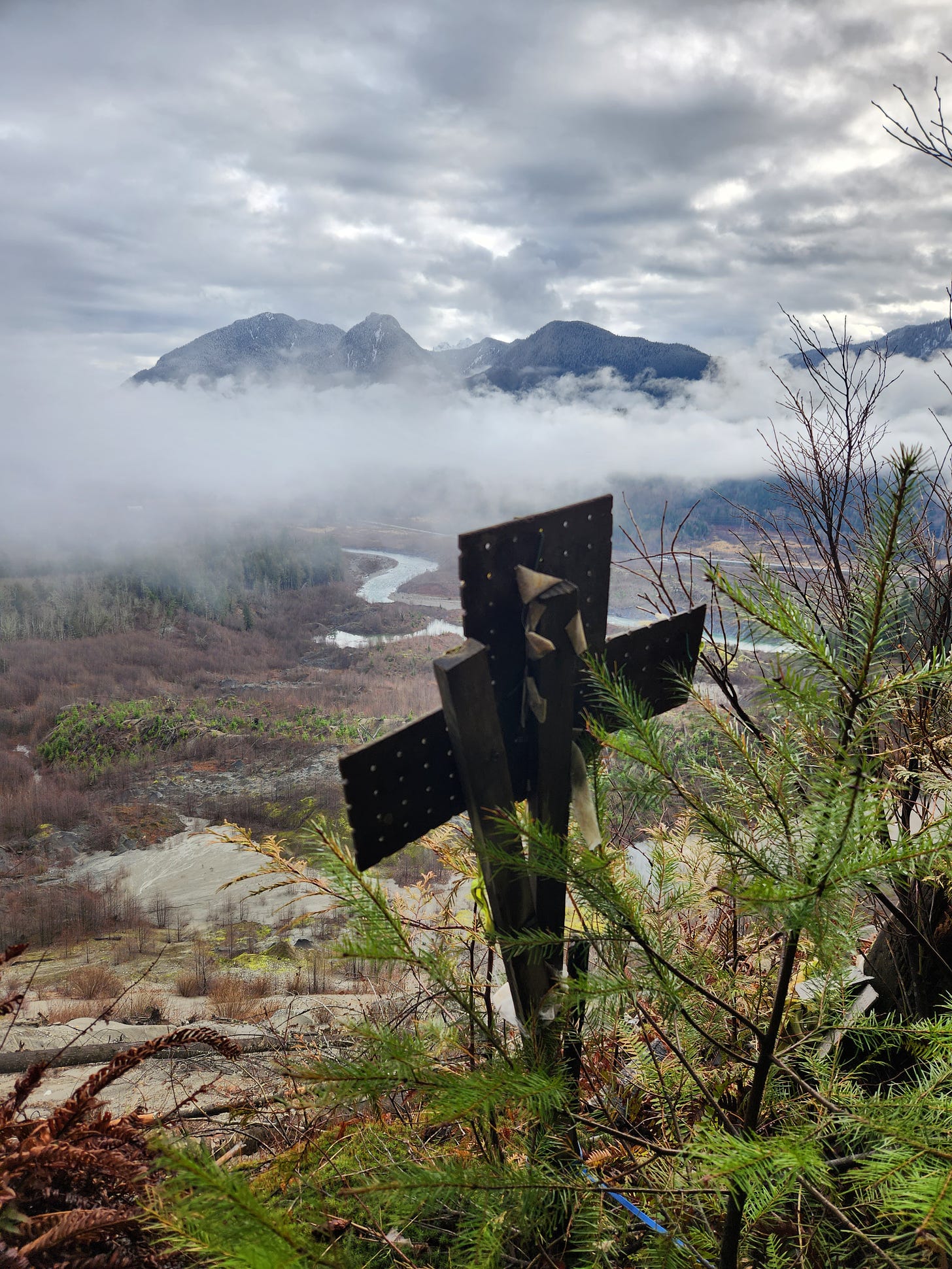 handmade cross on a dropoff before a river valley and mountain backdrop with clouds