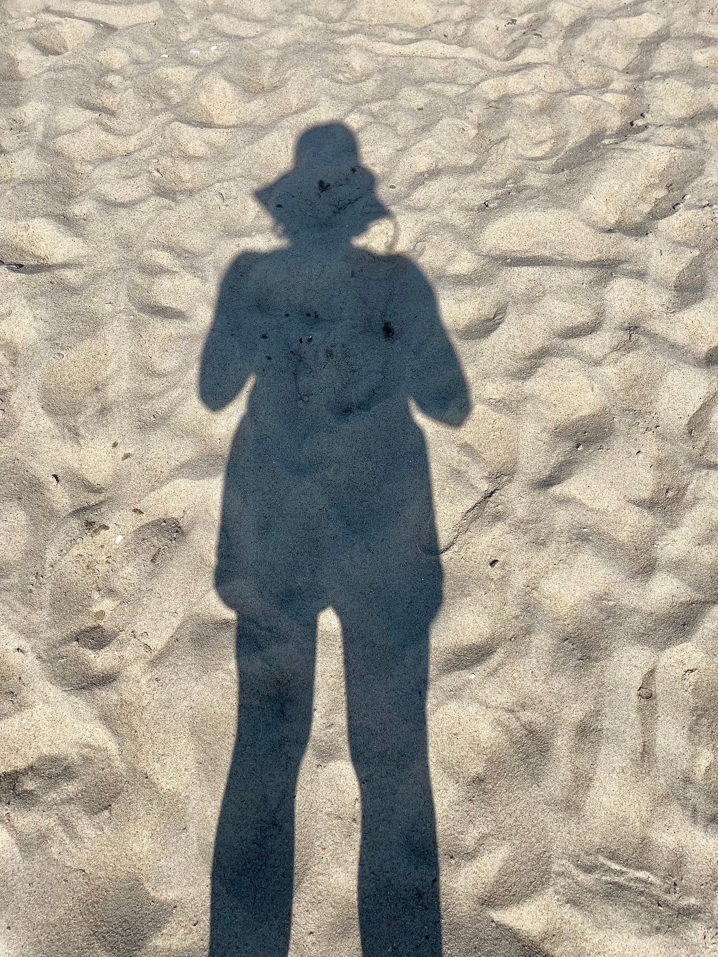 Long shadow of woman in wide-brimmed hat, on beach sand.