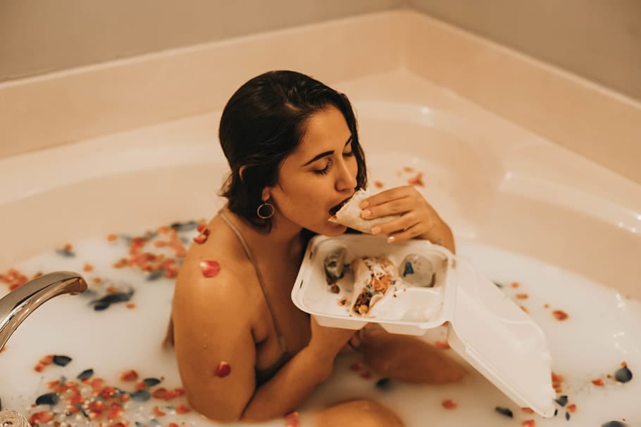 A woman in a large bathtub filled with milky white water and flower petals eating a burrito out of a takeout container.