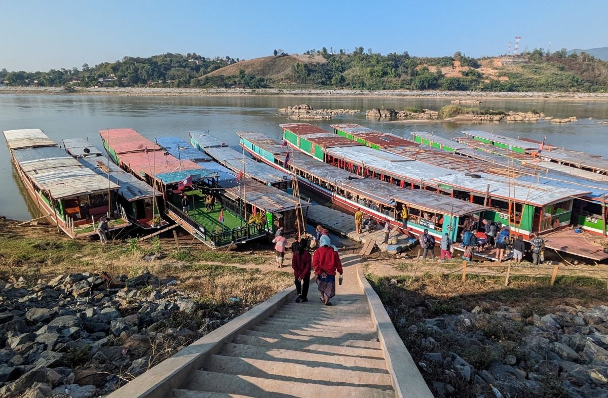 a line of long, narrow, low-slung boats, brightly colored, lined up on a river bank awaiting passengers, some of whom are walking down a long staircase toward them. The river and hills are in the background.