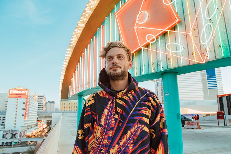 A person stands smiling in a vibrant, patterned jacket near a neon-lit sign with dice. In the background, there are buildings and the "FREMONT" sign.