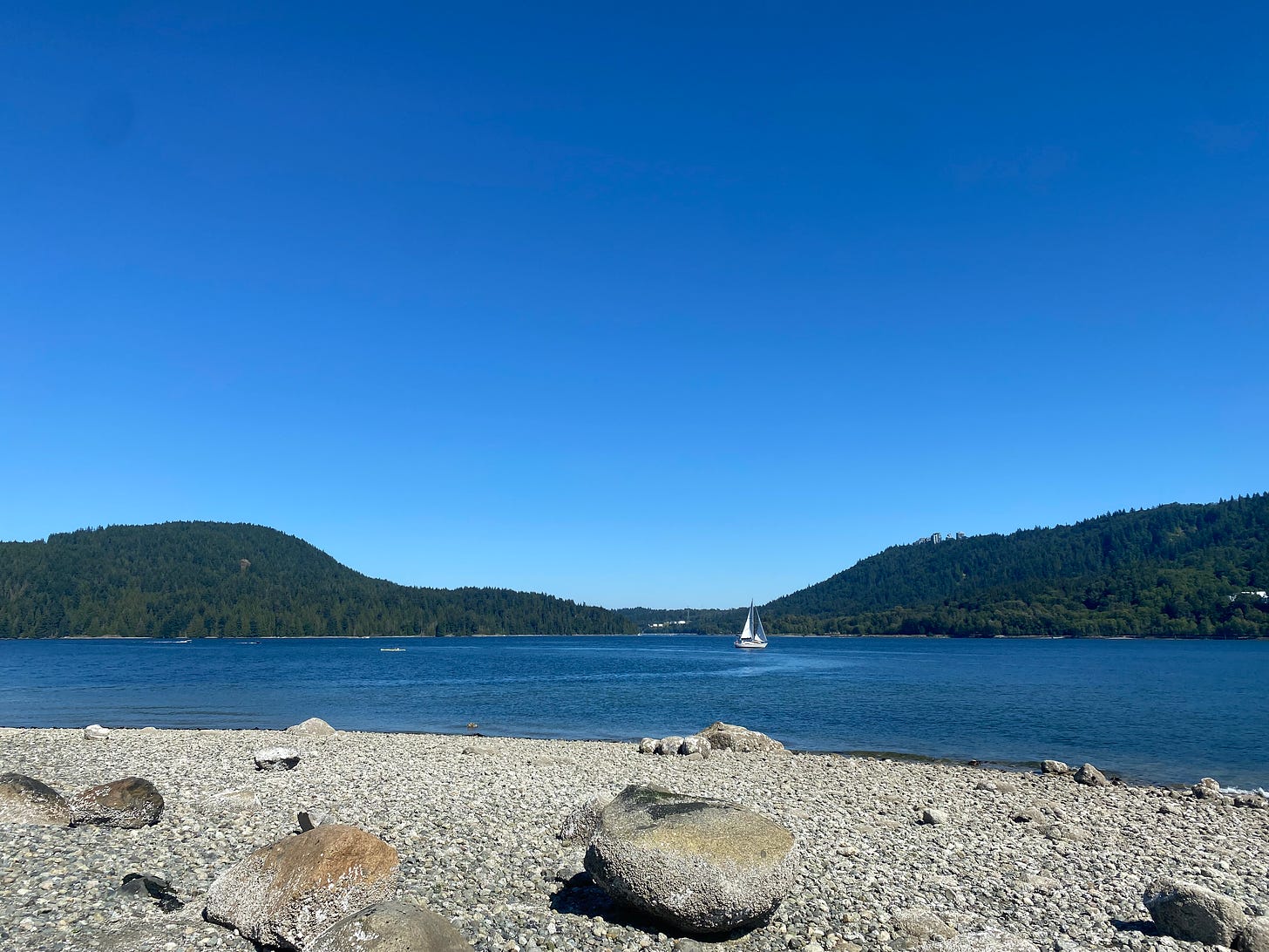 A view across the inlet from Cates Park, looking toward the greens of Burnaby and Port Moody along the horizon. A white sailboat sits alone near the centre of the frame, on the water between the two hills. The foreground shows a rocky beach with barnacles along the larger rocks. The water and the sky are the same shade of blue.