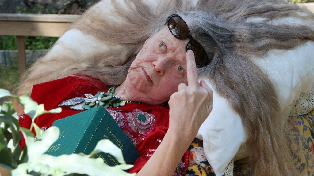 A disabled white woman is outside. Her grey hair is splayed out on a white pillow under head. She has sunglasses pushed up above her face and a book lies on her torso. She gives a middle finger to the camera with a severe look.