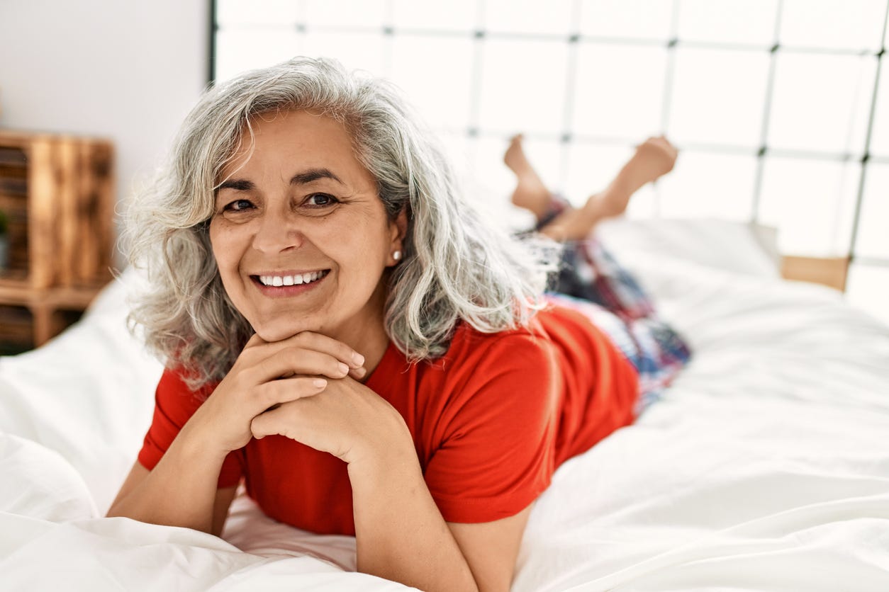 Smiling, grayed-haired Latina woman laying on bed on her stomach, propped up by her elbows. Wearing red blouse and plaid slacks. Her feet and legs out of focus in background.
