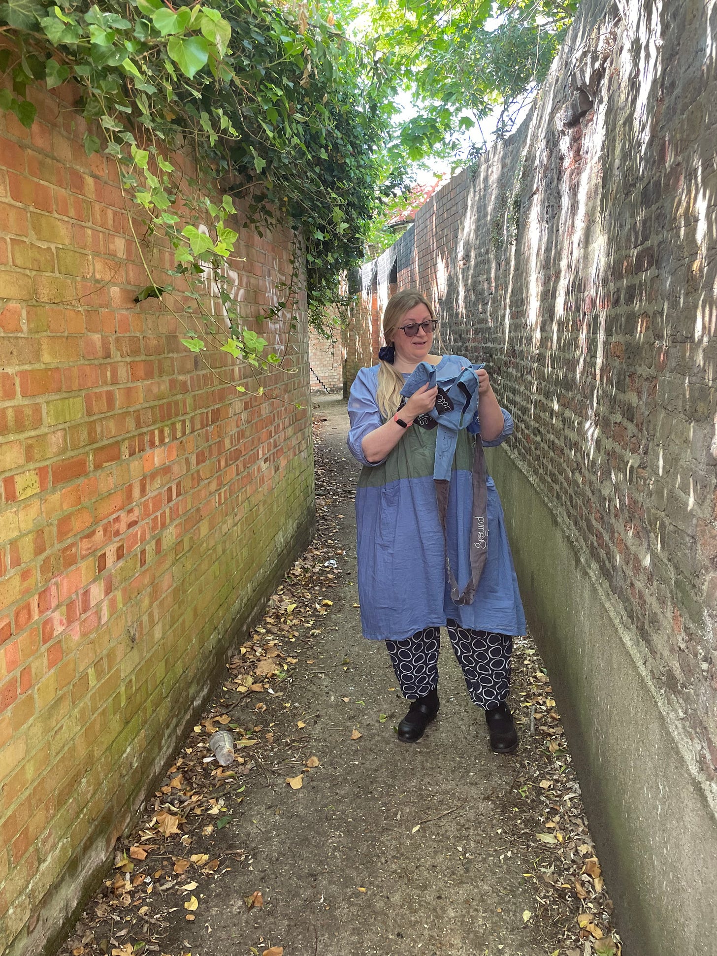 A white woman in a blue dress stands in an alleyway between two walls, holding strips of fabric
