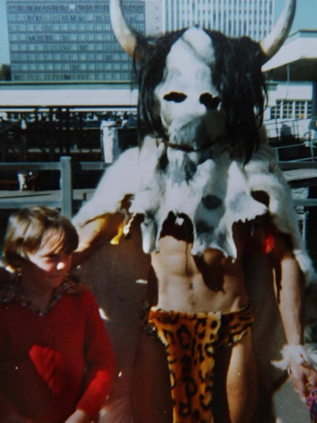 Photo that appears to be taken on a park or boardwalk, of a kid next to an adult. The adult is topless, but largely shrouded by a white animal skin that covers head as well as upper body. It has eyes cut out, black hair attached, and horns sticking out the side. He's also wearing a cheetah print loincloth