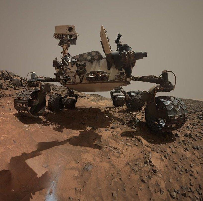 A low-angle color photo of the Curiosity rover, its six wheels resting on the rocky Martian surface, with a grey/brown sky behind it