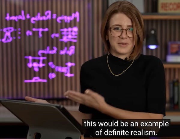 Screen capture from IBM's video about quantum entanglement. A woman who appears to be about 30, facing the camera, gestures with both hands to her right. On screen behind her there is some illegible backward writing that appears to be floating in air.. The caption of what she's saying is 'this would be an example of definite realism.' In fact the purple writing is on a piece of clear plexiglass in the set and has nothing to do with the current video. It's an incongruous juxtaposition.