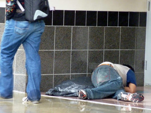 Photo of a homeless man sleeping in a doorway on a rainy day as another man passes by, captioned "Soylent green is people."