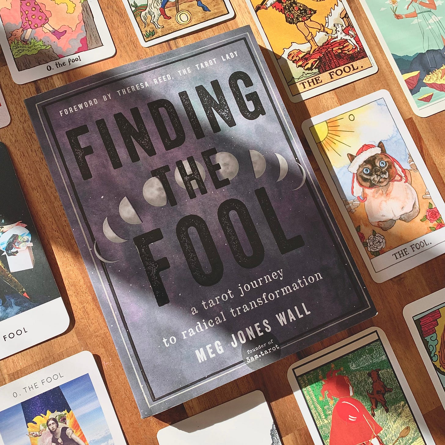 A paperback copy of Finding the Fool by Meg Jones Wall on a wooden tabletop, surrounded by different versions of the Fool tarot card