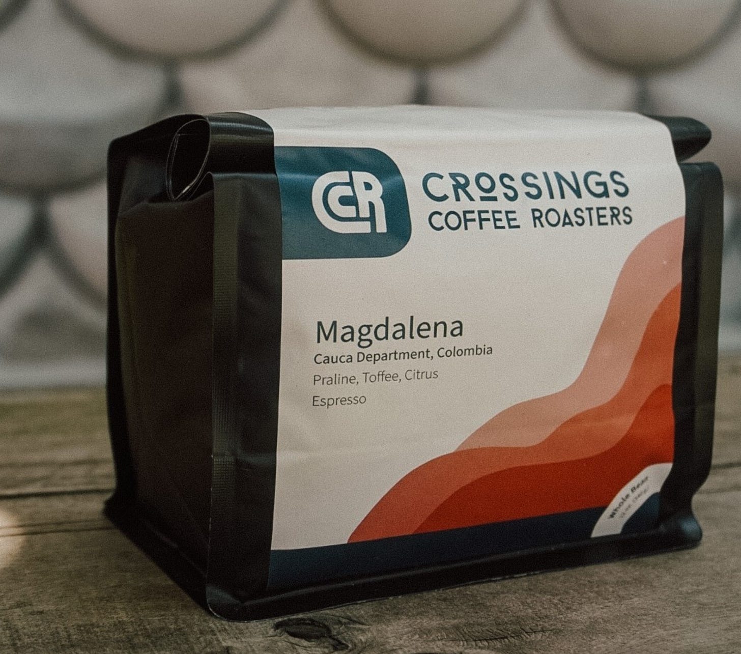 A close up of a black coffee bag with a white label and the Crossings Coffee Roasters typographic logo on it near the top of the bag.