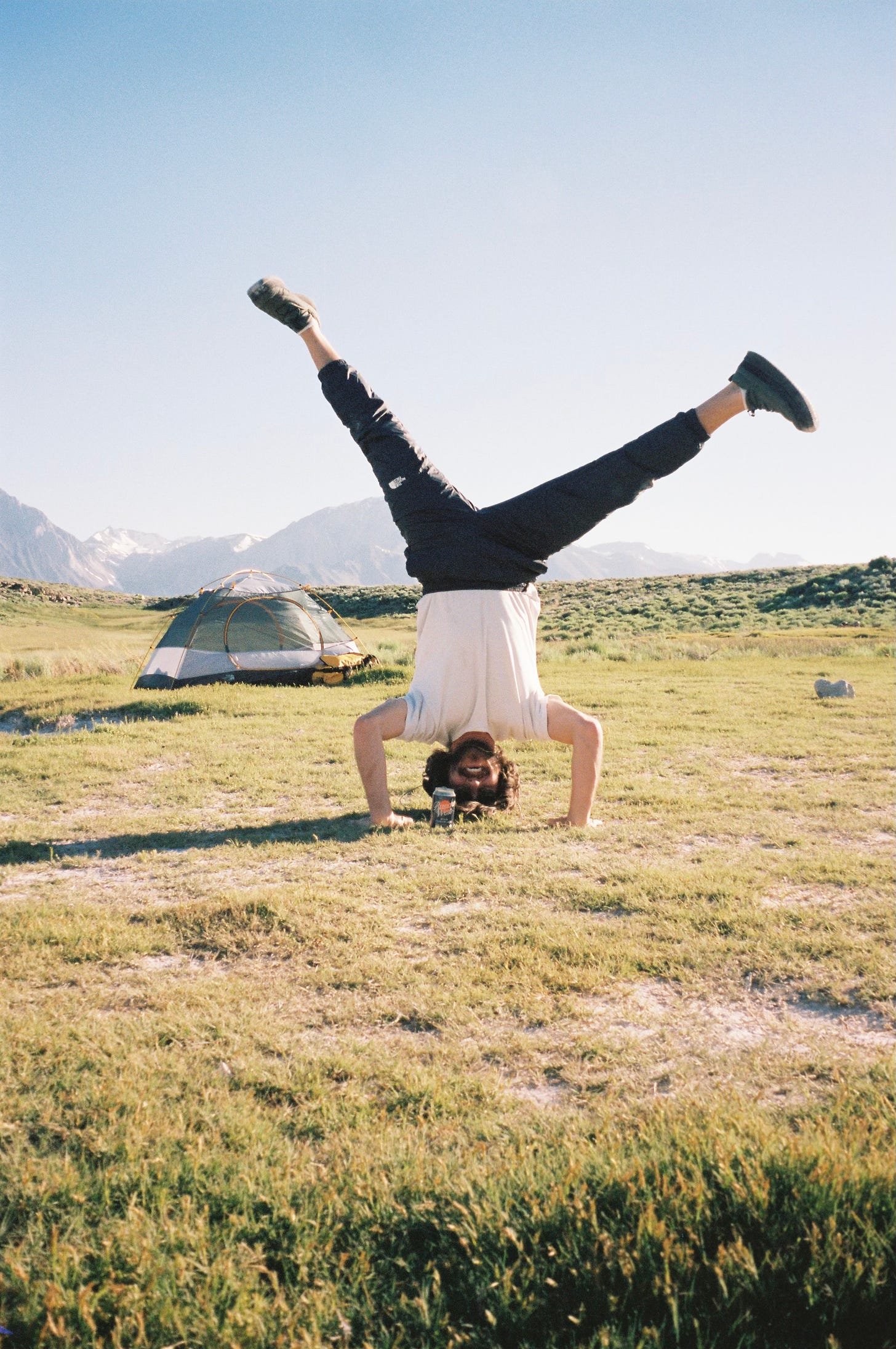 A man doing a headstand on a grassy hillside with a tent and mountains in the background.