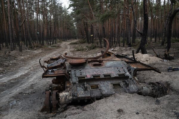 Rusty remains of a Russian tank are partly buried in snow and earth along a dirt road through a forest. 