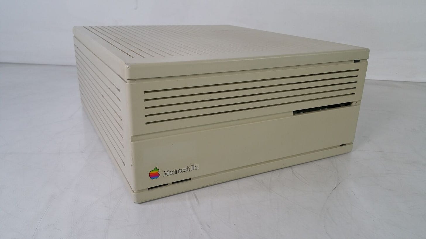 Mac IIci CPU. It was 5.5x11.9x14.4 inches. The front has a rainbow Apple logo, the inscription "Macintosh IIci", a floppy disk slot, and two little slots at the bottom where a programmer could add a little plastic "programmer's switch". It is sitting on a marble table.
