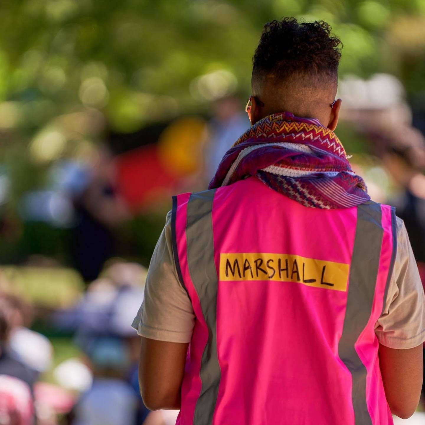 A photo of the back of a person wearing a pink hi-vis vest that reads 'marshall'. They are wearing a purple keffiyah and have short black curly hair with a fade and brown skin.