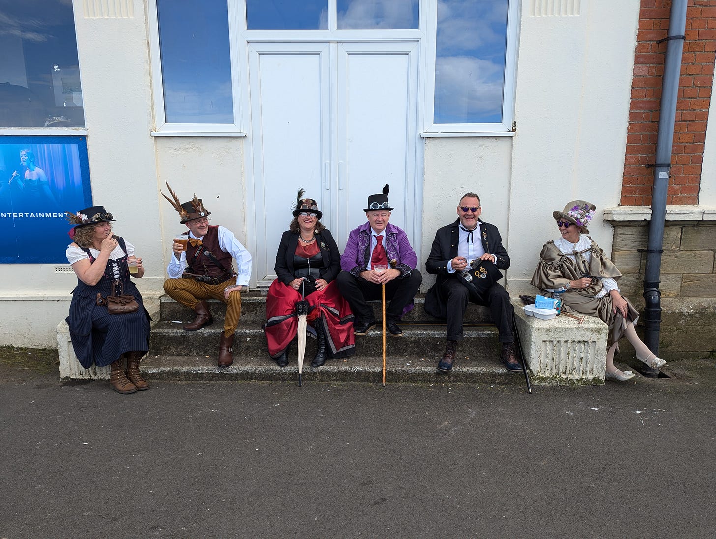 Six steampunkers sit outside, smiling at each other or the camera