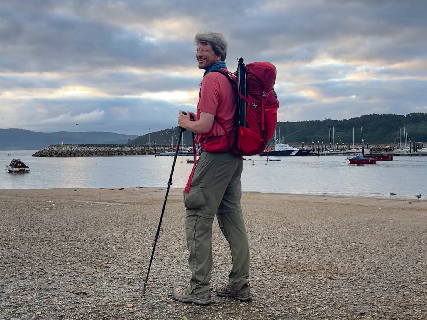 I'm standing at the harbor of Muxia, unshaven, with a red backpack, overlooking the water.