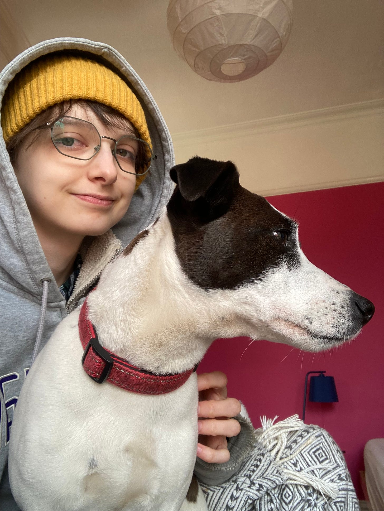 Selfie of Beck in a hoodie and yellow hat, scratching a brown and white spotted Jack Russell sitting on their lap but looking away from the camera, into the distance like there is a heavy weight on her mind. What horrors has she seen? We will never know. 