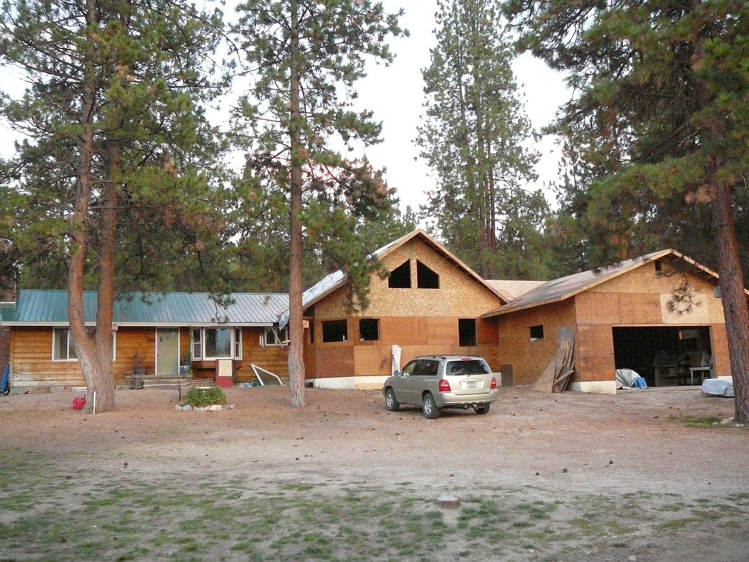 a small house with a larger, still under construction portion being added on, surrounded by pine trees