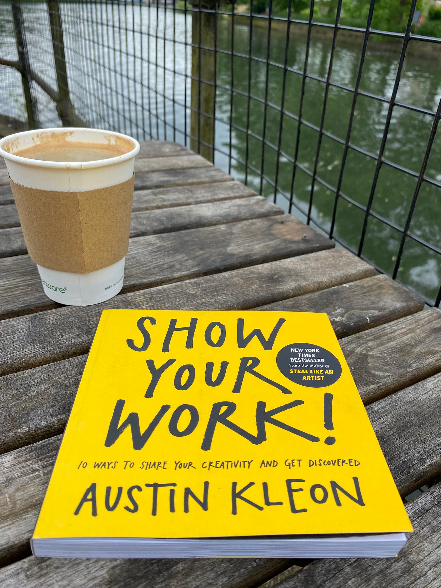 A copy of the book Show Your Work by Austin Kleon is laying on a garden table in front of a cup of coffee. The book is square with a bright yellow cover. In the background, behind a wire fence, is a river.