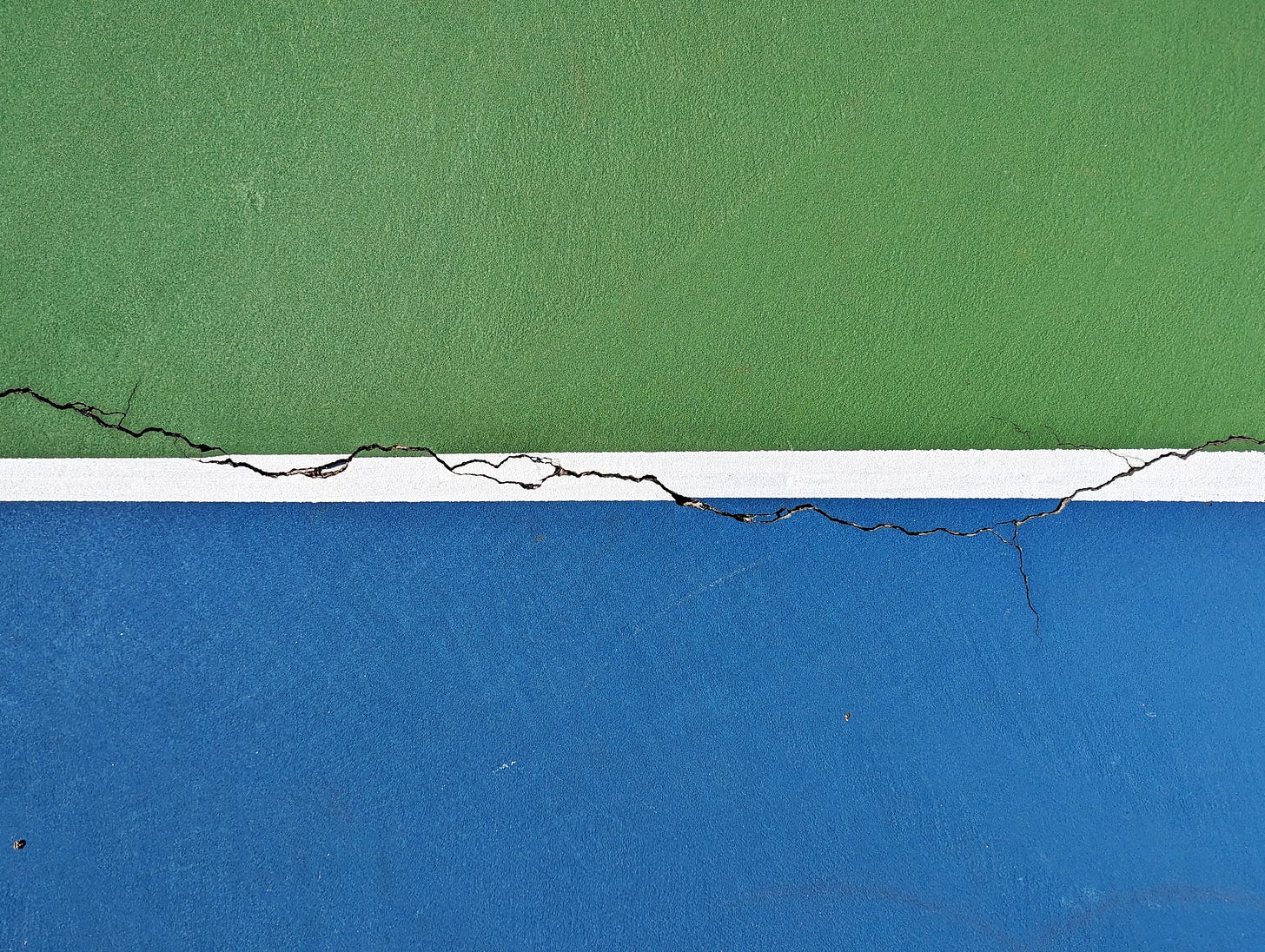 A detail shot of a tennis/pickleball court showing a crack along a horizontal white line dividing a blue area from a green area.